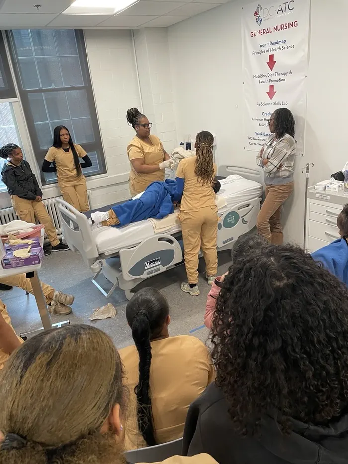 Apprentices in scrubs practising patient care around a hospital bed in a skills lab.