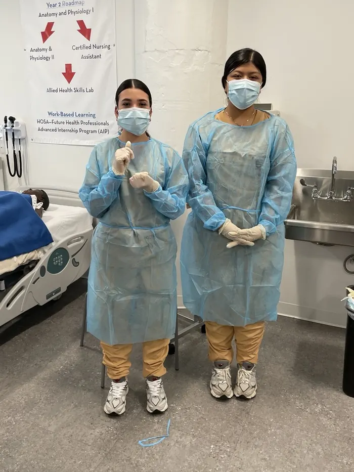 Two apprentices in gowns, masks and gloves standing beside a hospital bed in an allied health skills lab.