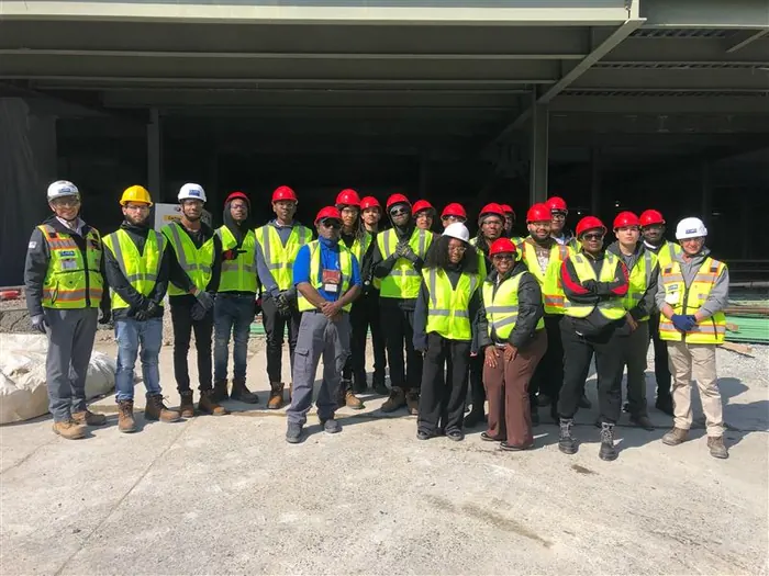 A large cohort of apprentices in high-visibility vests and hard hats posed together indoors.