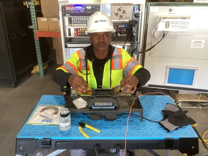 An apprentice seated at a fibre splicing bench in a hard hat and high-visibility vest.