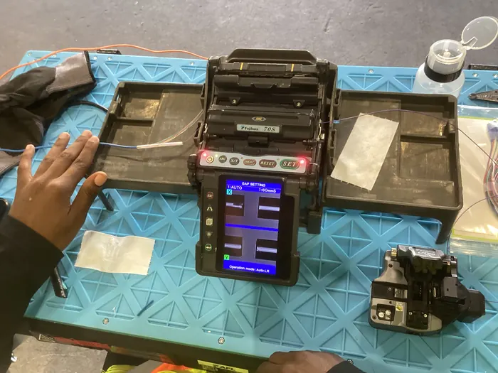 A fibre fusion splicer open on a workbench as an apprentice prepares a splice.