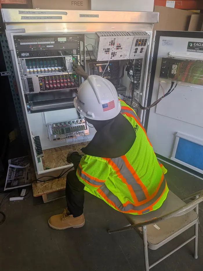 An apprentice in a hard hat and high-visibility vest wiring a control panel.