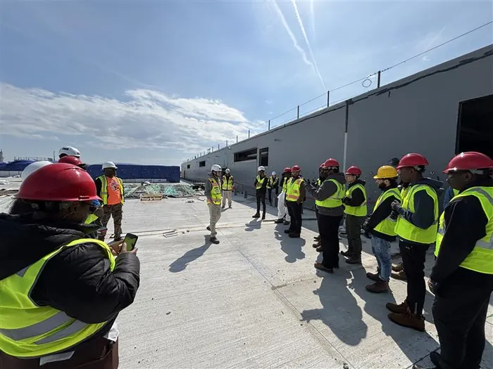 Apprentices in high-visibility vests and hard hats walking a construction site under an open sky.