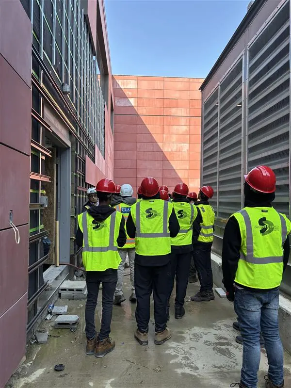 Apprentices in high-visibility vests and red hard hats gathered at a wall during a site walk.