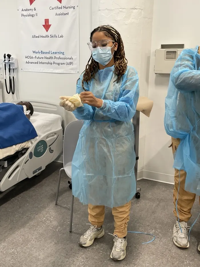 An apprentice in a gown, mask, goggles and gloves pulling on a second glove beside a hospital bed in an allied health skills lab.