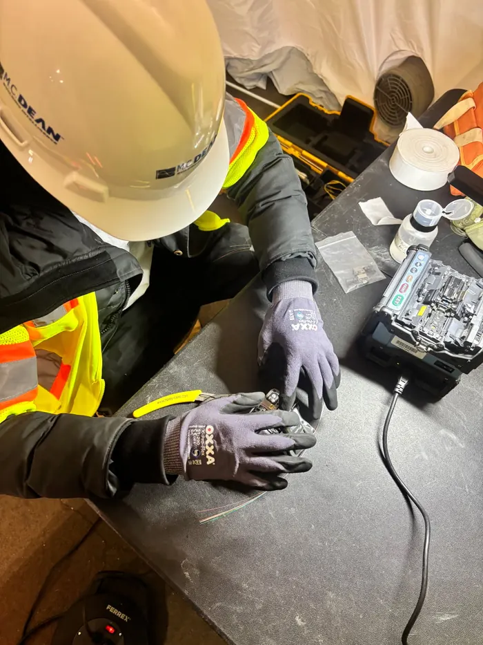 An apprentice in an MC Dean hard hat and gloves preparing fibre at a fusion splicer.