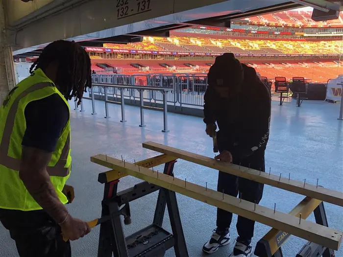 Two apprentices marking out a length of timber on sawhorses on a stadium concourse.